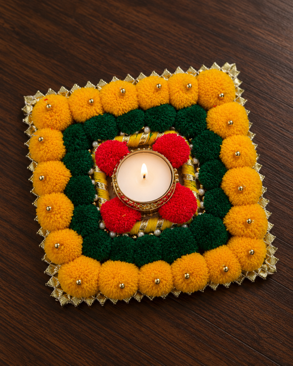 Close-up of the square rangoli piece with marigold-yellow and green pom-poms, gold beads and a lit tealight