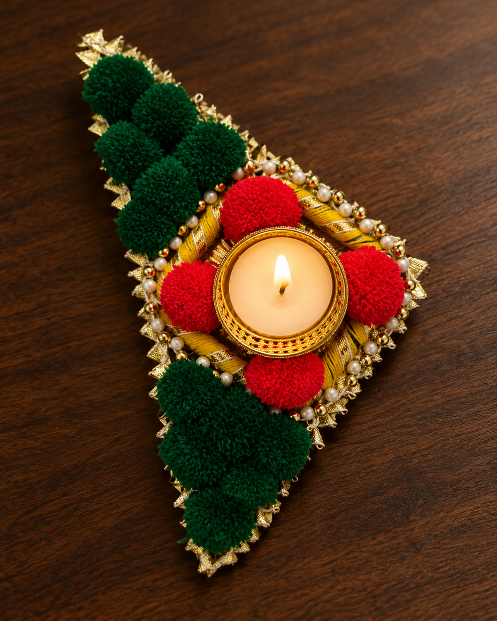 Close-up of a green triangle rangoli piece with red pom-poms, pearls, gota and a lit tealight